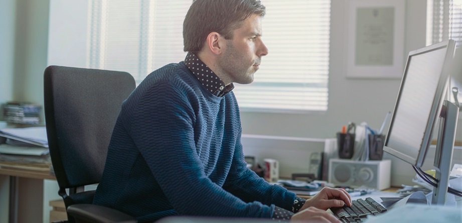 A imagem apresenta um cenário de home office, em que um homem está sentado à frente do seu computador. Ele olha para a tela e está teclando. Ele está de lado na cena.