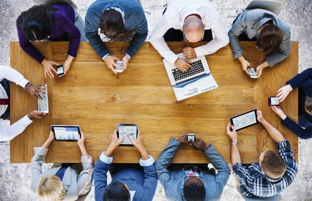 A imagem apresenta uma mesa vista de cima, com 10 pessoas em sua volta, aparentemente trabalhando. Cada uma usa um tipo diferente de hardware para trabalhar, como laptop, celular ou tablet. 