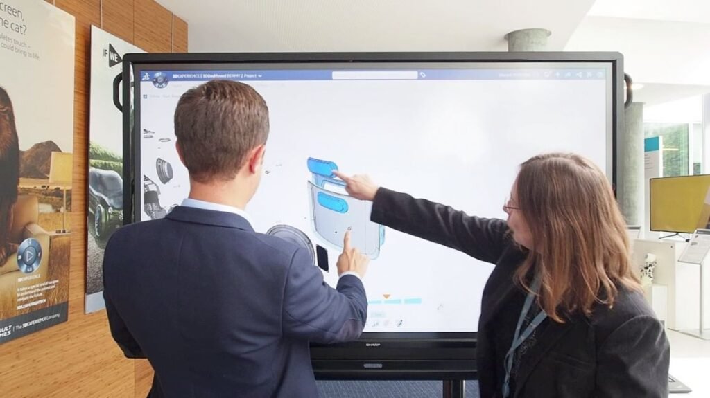 A imagem apresenta um homem e uma mulher, de costas, olhando para uma grande tela às suas frentes. A mulher está indicando algo na tela, o que aparece estar mostrando um software CAD.