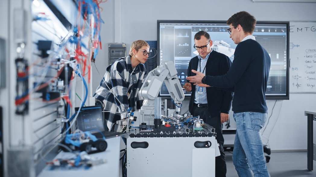 A imagem apresenta 3 profissionais, dois homens e uma mulher. Todos olham para uma mão robótica à sua frente. Eles estão em uma sala de tecnologia.