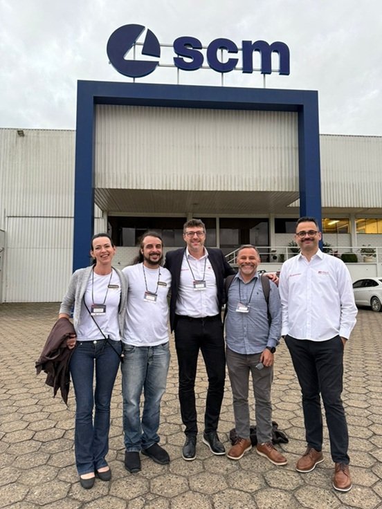 A imagem apresenta uma foto com 5 pessoas, sendo 2 da empresa SWOOD, posando em frente à empresa SCM. Há um logo na parte de cima da fachada da empresa.